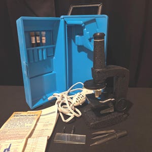 May include: A vintage black microscope with a blue carry case. The microscope has a light and is connected to a power cord. The case contains vials and a manual. Additional accessories are included.
