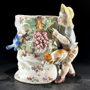 May include: A detailed porcelain vase featuring a young figure in a yellow hat and floral pants, pointing towards a bunch of grapes. A small dog jumps up, and a blue bird sits on the vase. The vase is decorated with leaves, flowers, and a textured surface.