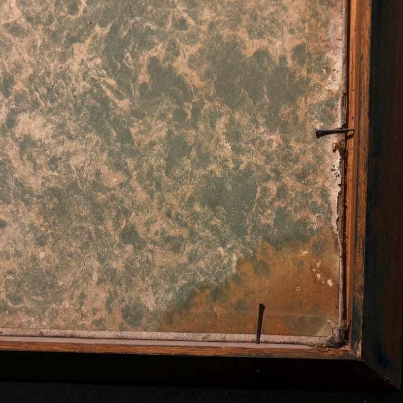 May include: Close-up of a framed artwork with a textured, mottled surface in shades of green and tan. The wooden frame is visible, with a nail securing the canvas. The artwork has a vintage aesthetic.