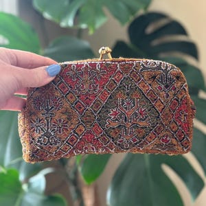 May include: A vintage beaded clutch with a gold clasp. The clutch features an intricate pattern in shades of red, brown, and gold. The beaded design creates a textured surface, adding to its vintage appeal. The clutch is held in a hand.