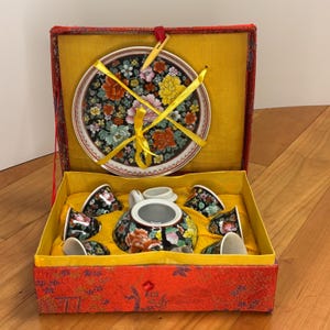 Puede incluir: Un juego de té vintage en una caja roja y amarilla. El juego incluye una tetera, seis tazas, un platillo y un plato. Las piezas de porcelana son negras con diseños florales coloridos. La caja es roja con interior amarillo.