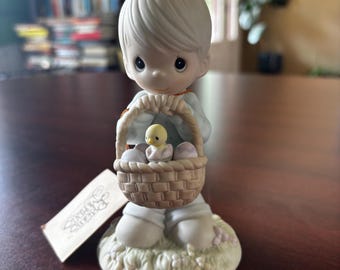May include: A Precious Moments figurine of a young person holding a basket with eggs and a chick. The figurine is light blue and white, with a tag that reads "Precious Moments". The figurine is on a grassy base.