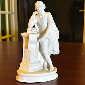 May include: A white porcelain figurine of a man in period clothing, leaning on a pedestal with books and a scroll. The figure is in a thoughtful pose, with one hand on his chin. The statue is on a round base.