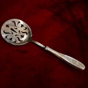 Może przedstawiać: Srebrna łyżka do serwowania z ażurową misą i długim uchwytem. Misa ma dekoracyjne, wycięte wzory. Uchwyt jest gładki i zaokrąglony, z detalicznym fragmentem w pobliżu misy. Łyżka jest umieszczona na ciemnoczerwonym tle.