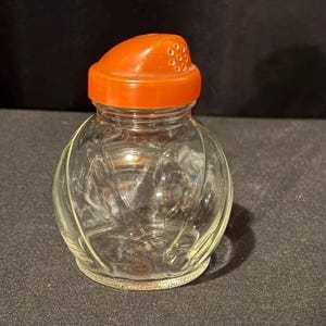 May include: A clear glass spice jar with an orange plastic lid. The jar has a rounded shape with vertical etched lines. The lid has small holes for dispensing. A vintage kitchenware item.
