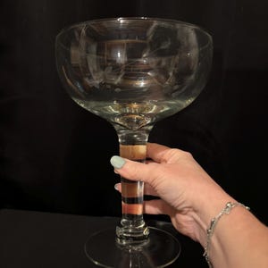 May include: A large, clear glass champagne coupe with a wide bowl and a tall, striped stem. The stem features bands of brown, tan, and orange. The glass has etched floral designs near the rim. The coupe sits on a round base.