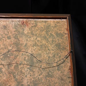 May include: Back of a framed artwork with a wooden frame. The artwork has a mottled green and tan background. A black wire is strung across the back for hanging. The frame is secured with small nails.