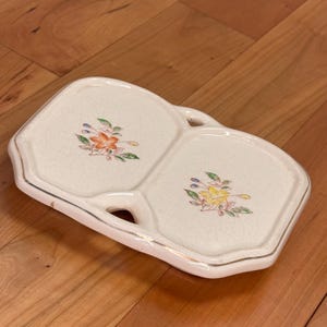 May include: Cream-colored, divided serving dish with floral designs. The dish has two compartments and a handle. The floral designs feature orange, yellow, and blue flowers with green leaves. The rim is trimmed with a gold-colored line.