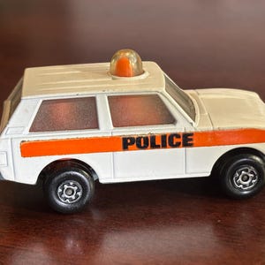May include: A white toy police car with black wheels and an orange stripe. The word "POLICE" is printed in black on the side. An orange light is on top of the car.
