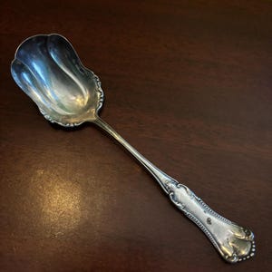 May include: A silver serving spoon with a wide, scalloped bowl and a long, slender handle. The handle features ornate detailing and a rounded end. The spoon is designed for serving and has a classic, elegant appearance.