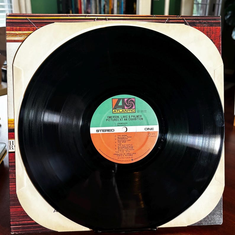 Op de afbeelding: Een vintage vinylalbum van Emerson, Lake & Palmer, getiteld "Pictures at an Exhibition". De zwarte plaat heeft een kleurrijk label met het Atlantic Records-logo. De albumhoes is zichtbaar en toont een bruin en wit ontwerp.