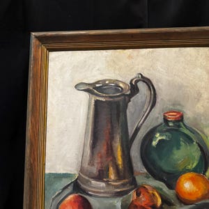 May include: Oil painting of a still life with a silver pitcher, a green bottle, and oranges. The painting is framed in a wooden frame. The background is a light beige color.