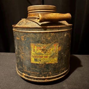 May include: An antique metal container with a wooden handle and a label. The container is dark blue with rust and wear. The label has text in red and yellow. The handle is a light brown color.