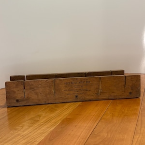 May include: A vintage wooden miter box, likely for woodworking. The box is rectangular with angled slots for precise cuts. The text "ROCK MAPLE MITER BOX" is visible on the front. The wood has a warm, aged tone, suggesting it's been used.