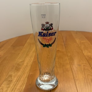 Puede incluir: Una copa de cerveza transparente con las palabras "Kaiser Weisse" en escritura azul y roja. La copa tiene un escudo decorativo y una marca de medición de 12,0. La copa está sobre una superficie de madera.