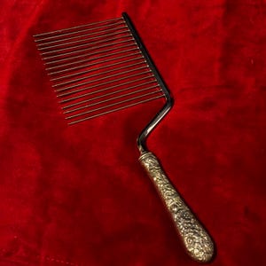 Op de afbeelding: Een zilveren taartsnijder met een reeks parallelle draden om te snijden. De handgreep is versierd met een bloemmotief. De snijder staat tegen een rode achtergrond.