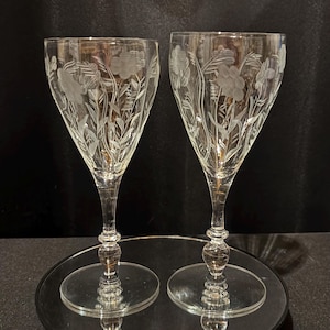 Puede incluir: Dos copas de vino transparentes con diseños florales grabados. Las copas tienen un tallo delgado y una copa ancha, y el patrón floral incluye flores y hojas. Las copas se exhiben sobre una superficie espejada.