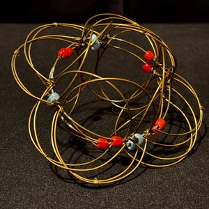 May include: A handcrafted, abstract sculpture made of intertwined gold-colored wire. The sculpture features red and light blue beads at various connection points, creating a unique and artistic design. The piece is displayed against a dark background.