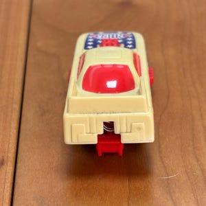 Op de afbeelding: Een vintage speelgoedauto in cr&egrave;me en rood. De auto heeft een rode cabine en een cr&egrave;me carrosserie met het woord "NHRA" en sterdetails. De speelgoedauto heeft een veermechanisme en is gemaakt van plastic.