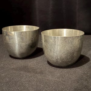 May include: Two small, silver-colored metal bowls with a rounded, cup-like shape. The bowls have a slightly textured surface and a simple, minimalist design. The bowls are likely vintage or antique.