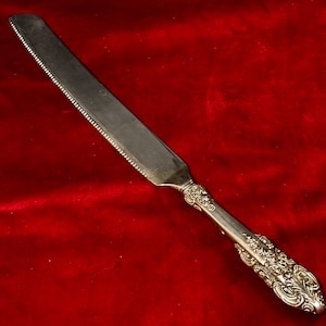 May include: A silver-colored knife with a serrated edge and ornate handle. The knife is displayed against a red background. The handle features detailed floral and scrollwork designs, adding a touch of elegance to the utensil.
