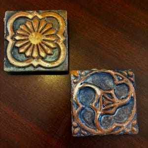 Può includere: Due piastrelle quadrate in ceramica con intricati disegni floreali. Una piastrella presenta un motivo a raggiera nei toni dell'arancione e del giallo, mentre l'altra mostra un disegno geometrico con accenti arancioni e blu. Le piastrelle hanno un aspetto rustico e artigianale.