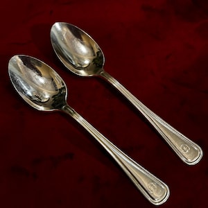 Puede incluir: Dos cucharas plateadas con superficie reflectante se muestran sobre un fondo rojo oscuro. Las cucharas tienen un diseño clásico con un mango detallado y un cuenco redondeado. Las cucharas están en ángulo, mostrando su elegante forma.