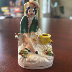May include: A vintage porcelain figurine of a seated figure in a green jacket and hat, holding a book. The figurine has a yellow pot and is decorated with colorful floral accents. The base is white with a cream-colored glaze.
