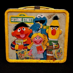 Pode incluir: Lancheira vintage de metal amarelo da Vila Sésamo com personagens como Monstro das Bolachas, Bert e Ernie. A lancheira tem uma pega amarela e um gráfico colorido do logótipo e personagens da Vila Sésamo. Um item colecionável nostálgico.
