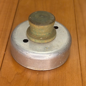 May include: A round metal biscuit cutter featuring a weathered, light green wooden knob handle centered on top. The silver-toned metal base has a smooth, cylindrical wall with two small circular ventilation holes positioned near the handle.