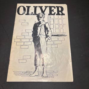 May include: Vintage paperback book cover featuring the title "OLIVER" in bold black letters. The cover illustration depicts a young person in a long coat, standing in front of a brick wall and window, all in black and white.
