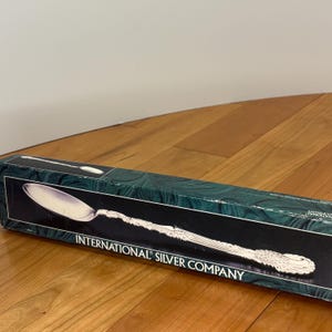 Op de afbeelding: Een zilveren serveerlepel in een doos van de International Silver Company. De lepel heeft een gedetailleerde steel en een grote, ovale kom. De doos is donkergroen met een zwart interieur en de bedrijfsnaam in het wit.