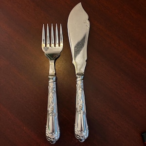May include: Silverware set featuring a fork and a butter knife. The fork has four tines, and the butter knife has a curved blade. Both utensils have ornate, detailed handles with a floral design. The set is made of a shiny, reflective metal.