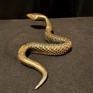 May include: A detailed bronze-colored snake sculpture with a textured body and a curved posture. The snake's head is raised, and its scales are intricately carved. The sculpture is set against a dark background, highlighting its metallic sheen.