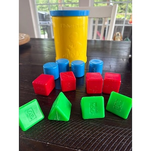 May include: A yellow Fisher-Price plastic canister with a blue lid, accompanied by a collection of colorful plastic shapes including blue cylinders, red cubes, and green triangular prisms, all featuring embossed details on their surfaces.