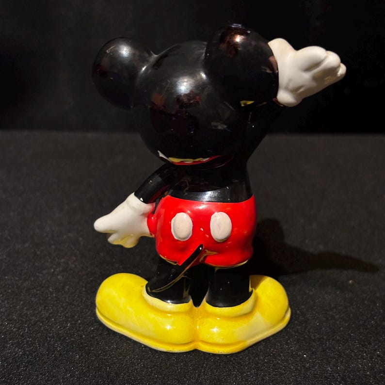 May include: A ceramic figurine of Mickey Mouse in a classic pose. The figure features a black body, red shorts with white buttons, yellow shoes, and white gloves. The figurine is posed with one arm raised.