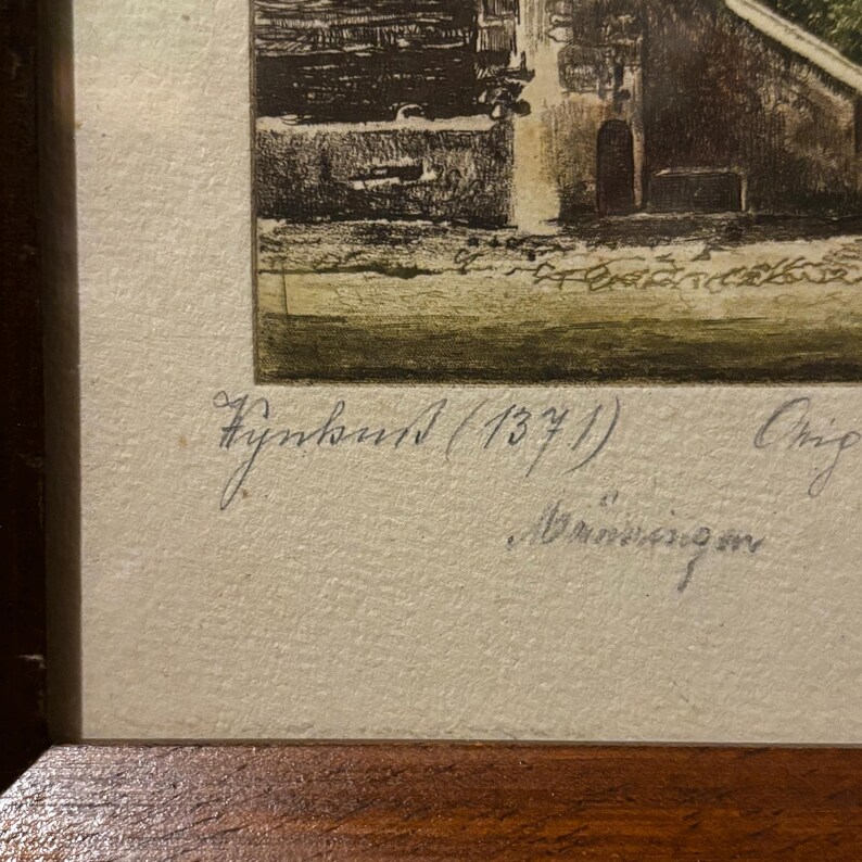 May include: A framed print depicting a detailed architectural scene, with a building featuring an arched doorway and surrounding landscape. The print is in a wooden frame. Handwritten text is visible.