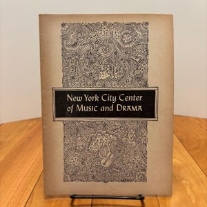 May include: A vintage program cover with the text "New York City Center of Music and DRAMA" in a black rectangle. The cover is tan with an intricate black and white floral and musical instrument design.