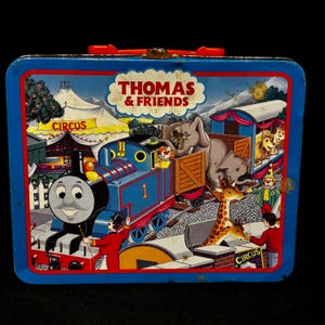 May include: Vintage Thomas & Friends lunchbox with a blue exterior and red handle. The front features a colorful illustration of Thomas the Tank Engine, circus animals, and the text "Thomas & Friends". The lunchbox has a metal clasp.