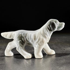 Puede incluir: Figura de cerámica de un perro en posición de caminar. El perro es principalmente blanco con marcas grises en la espalda y las orejas. La figura tiene un acabado brillante y está sobre un fondo oscuro.