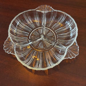 Peut inclure: Plat de service en verre transparent à trois compartiments et bord festonné. Le plat présente un motif radial avec des lignes verticales émanant du centre. Le plat est probablement utilisé pour servir des collations ou des apéritifs.