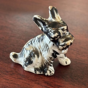 May include: A ceramic figurine of a rabbit in a sitting position. The rabbit is primarily white with black markings, and has its ears perked up. The figurine is on a dark brown wooden surface.