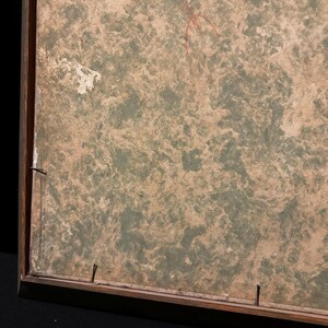 May include: A framed artwork with a textured, mottled surface in shades of green and brown. The wooden frame is visible, with nails securing the artwork. A red line is drawn on the surface.