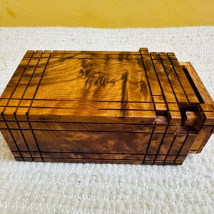Puede incluir: Una caja rectangular de madera con tapa corredera, hecha de una madera rica y cálida. La caja presenta una serie de ranuras paralelas y una tapa parcialmente abierta, revelando el interior. La veta de la madera es visible.