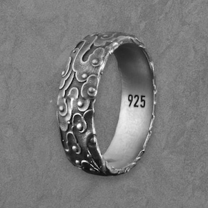 May include: A silver ring with a detailed, swirling design. The ring has a textured surface and the number "925" is stamped inside. The ring is shown on a grey surface.