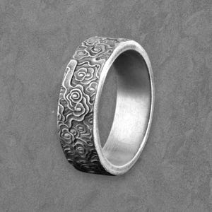 May include: A silver ring with a detailed floral pattern around the band. The ring is set against a grey background, highlighting the intricate design. The ring's interior is smooth and polished.