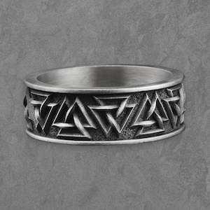 May include: A silver-colored ring with a geometric pattern of interlocking triangles and other shapes. The ring has a wide band with the design wrapping around the entire circumference. The ring is likely made of metal and has a slightly aged appearance.