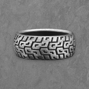 Op de afbeelding: Een zilveren ring met een geometrisch patroon. De ring heeft een brede band met een herhalend ontwerp van onderling verbonden vormen. De binnenkant van de ring is donker, wat contrasteert met de heldere zilveren buitenkant. De ring wordt op een grijze achtergrond getoond.