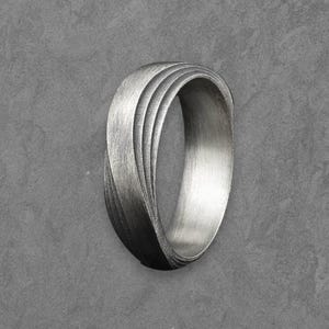 May include: A silver ring with a unique, layered design. The ring has a brushed metal finish and a curved, flowing shape. The design features multiple parallel lines that create a textured surface. The ring is a piece of jewelry.