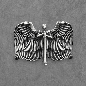 May include: A silver ring depicting an angel with large, detailed wings and a sword. The angel is the central focus, with intricate feather detailing. The ring has a textured, metallic appearance.